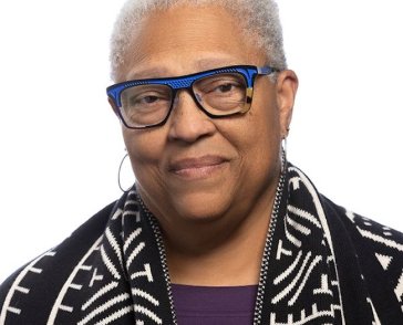 headshot of a woman with brown skin, close cropped gray hair and electric blue glasses frames. 